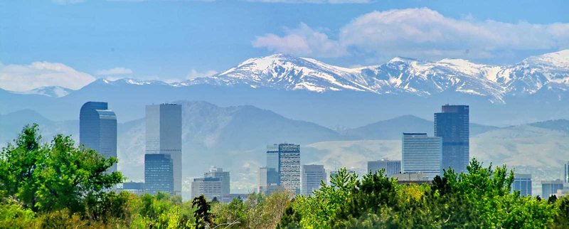 Denver skyline with trees, the buildings, and snow-capped mountains in the background.